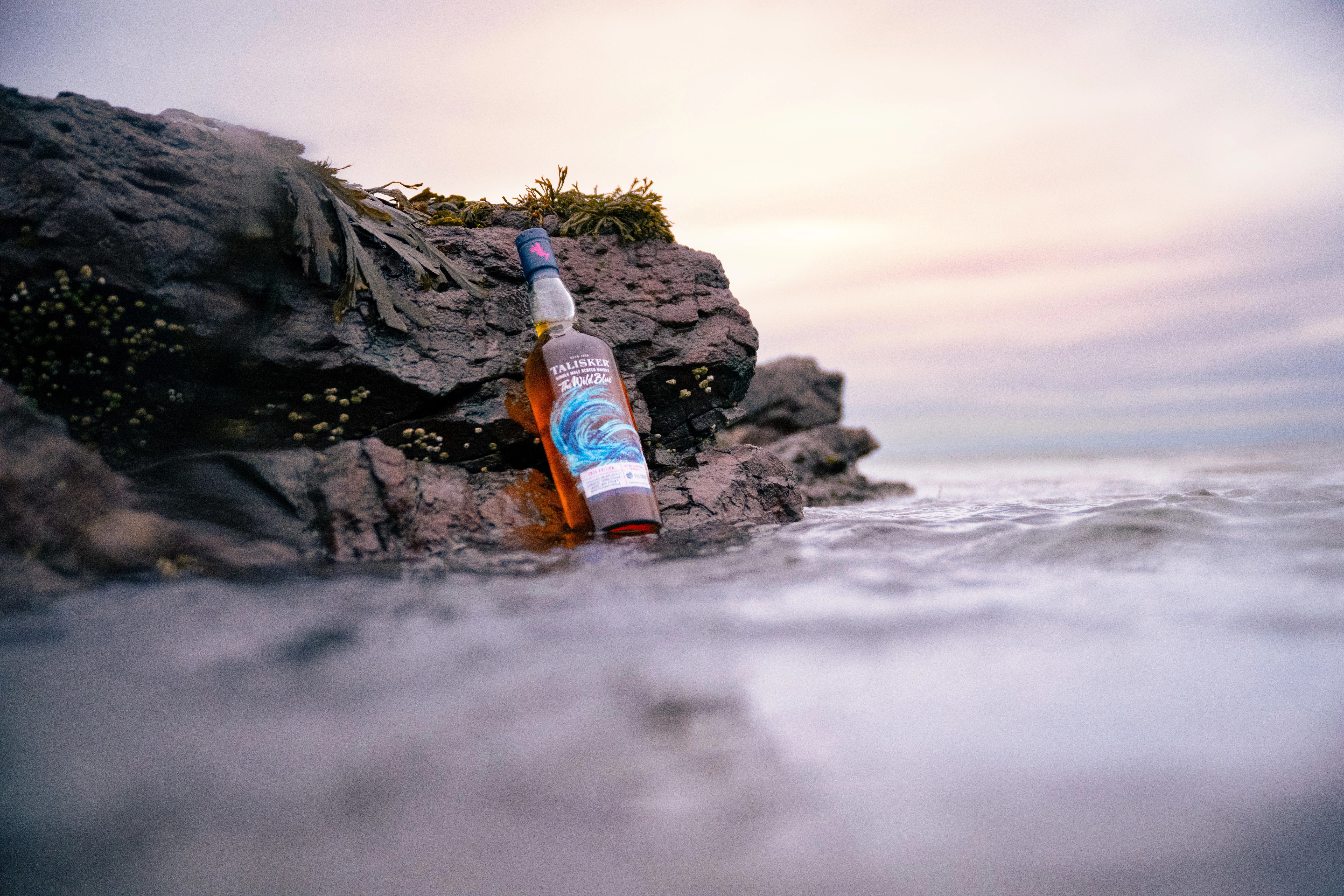 A bottle of Talisker The Wild Blue sits on the rocky shoreline as the sea sprays in the background