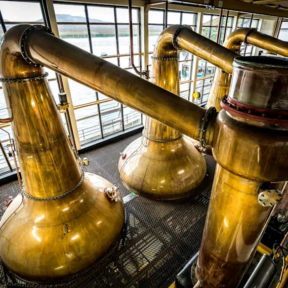 Several metal whisky stills are seen from above, sitting in front of floor to ceiling windows which overlook a body of water.