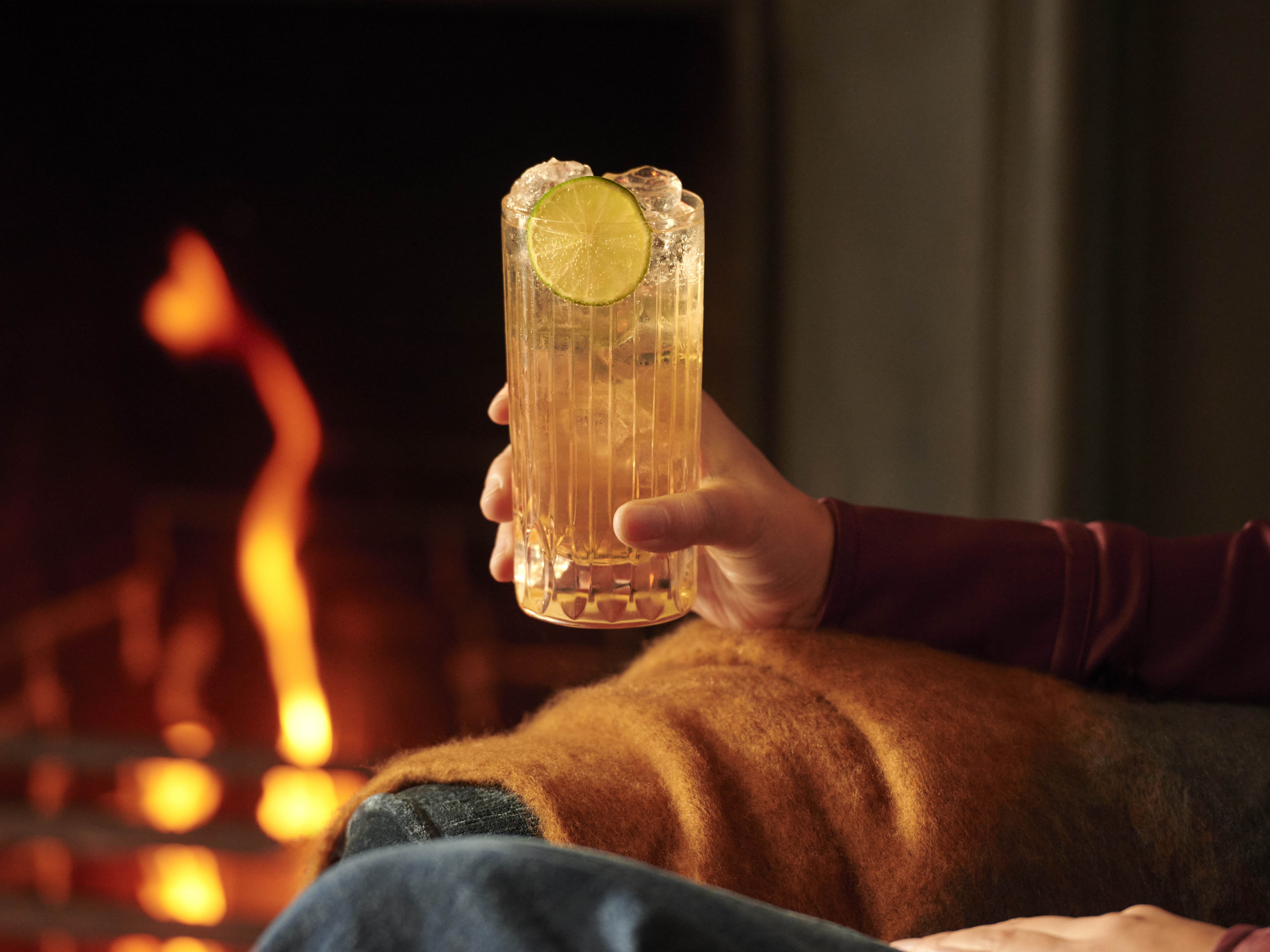 A person sits in an armchair by a fire, holding a whisky highball cocktail