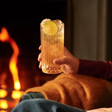 A person sits in an armchair by a fire, holding a whisky highball cocktail
