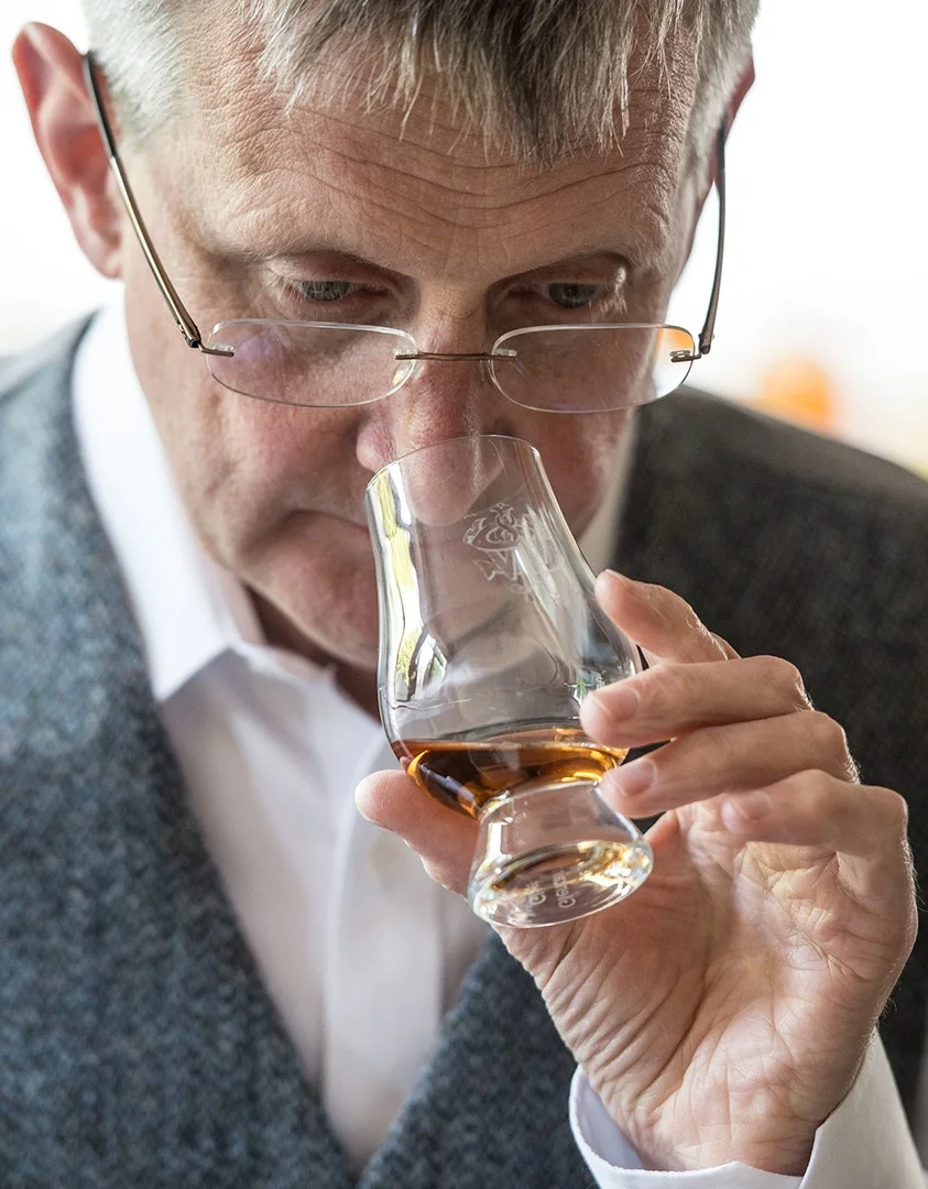 An older man smells a small glass of whisky.