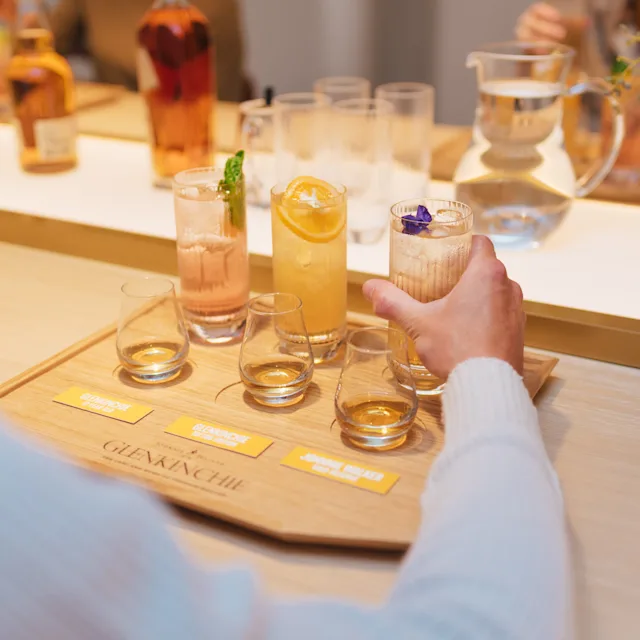 A wooden board engraved with the word ‘Glenkinchie’ sits on a table. On the board sits three dram glasses and three highball glasses, all containing an assortment of whisky and cocktails. A person place a hand round one highball glass.