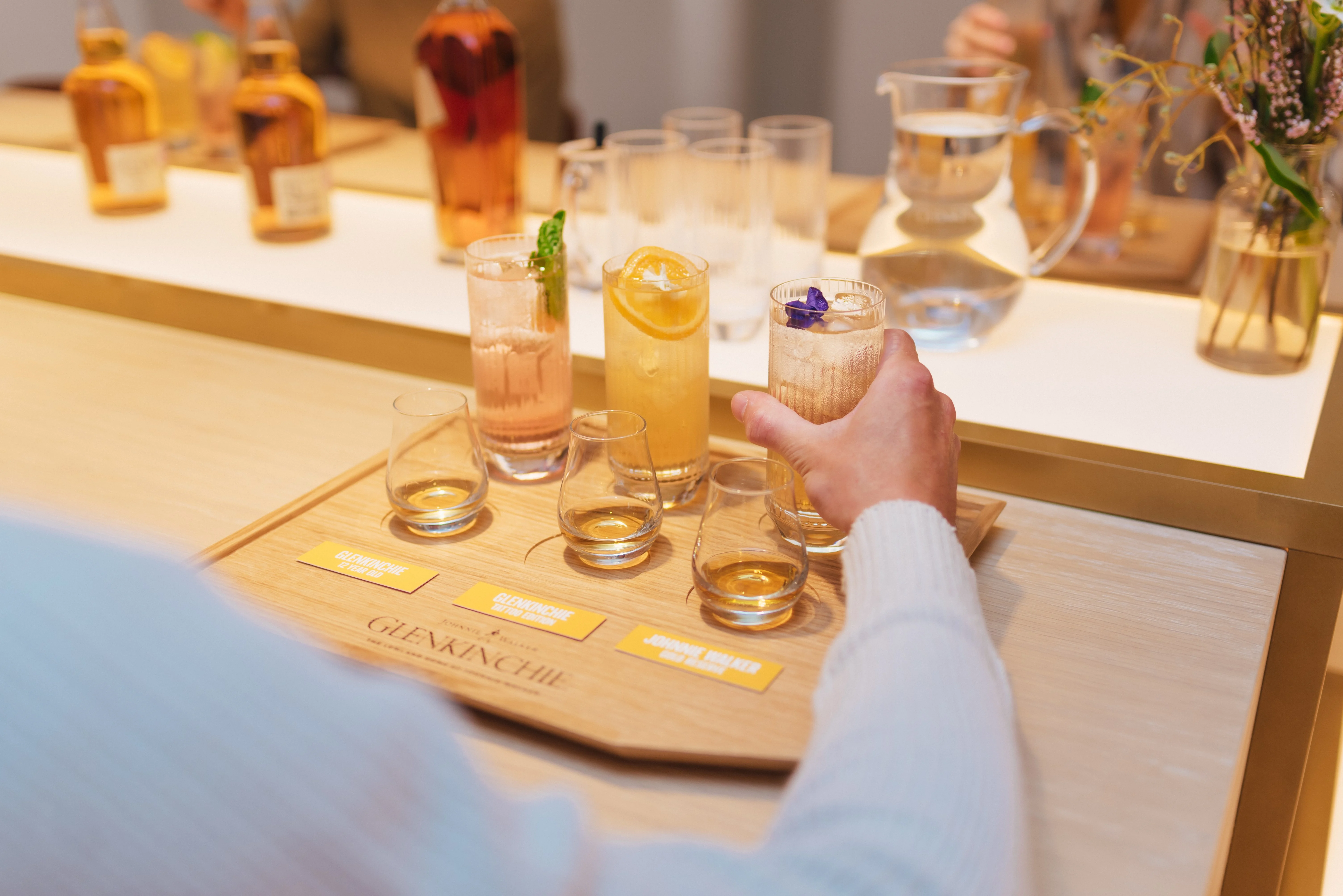 A wooden board engraved with the word ‘Glenkinchie’ sits on a table. On the board sits three dram glasses and three highball glasses, all containing an assortment of whisky and cocktails. A person place a hand round one highball glass.