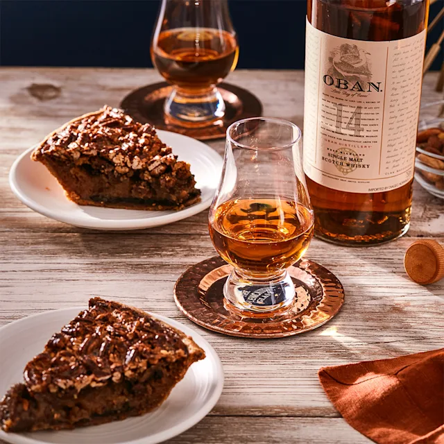 A bottle of Oban 14 year old sits on a wooden surface, next to two dram glasses filled with whisky and two plates of pecan pie.