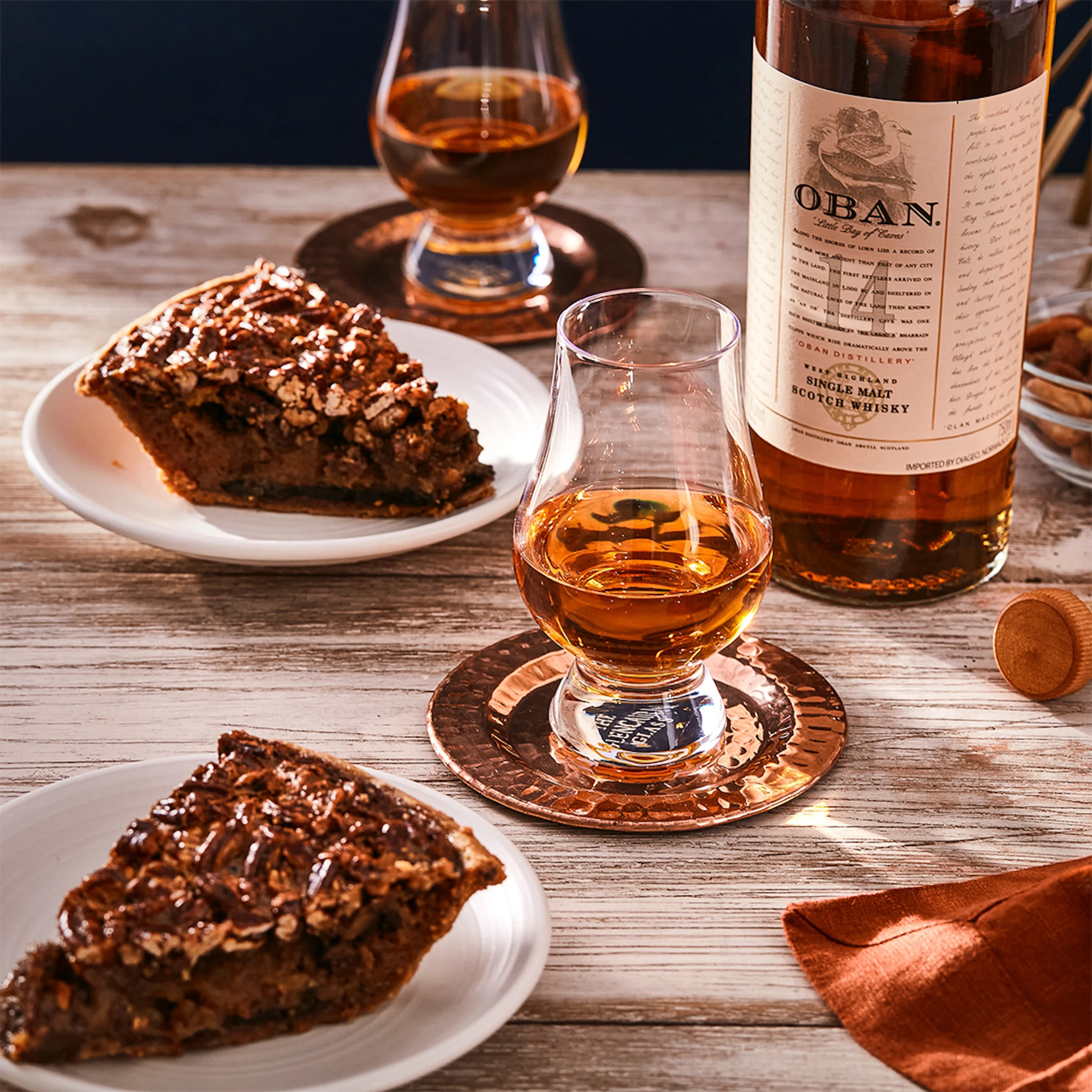 A bottle of Oban 14 year old sits on a wooden surface, next to two dram glasses filled with whisky and two plates of pecan pie.