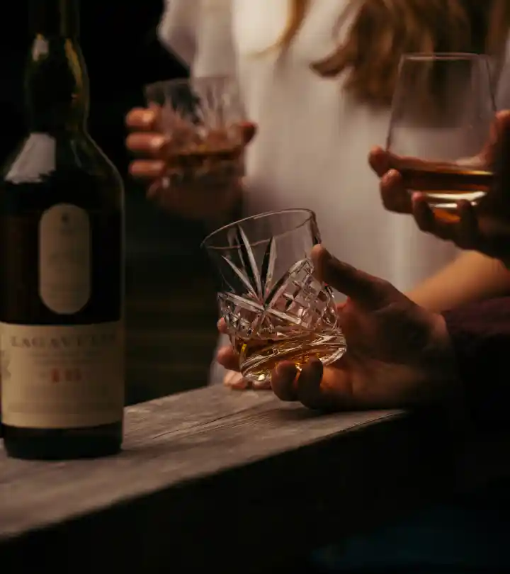 A bottle of Lagavulin whisky sits on a wooden table. Behind the bottle stands a person in white who holds a rocks glass containing whisky. In front of this person are two separate hands, each holding a rocks glass containing whisky.