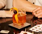 An Old Fashioned cocktail in a rocks glass sits on a glass table, surrounded by playing cards, dominos and some canapes. Two people sit behind the cocktail.