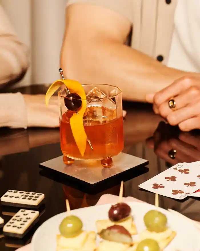 Ein Old-Fashioned-Cocktail in einem Rocks-Glas steht auf einem Glastisch, umgeben von Spielkarten, Dominosteinen und einigen Häppchen. Zwei Personen sitzen hinter dem Cocktail.