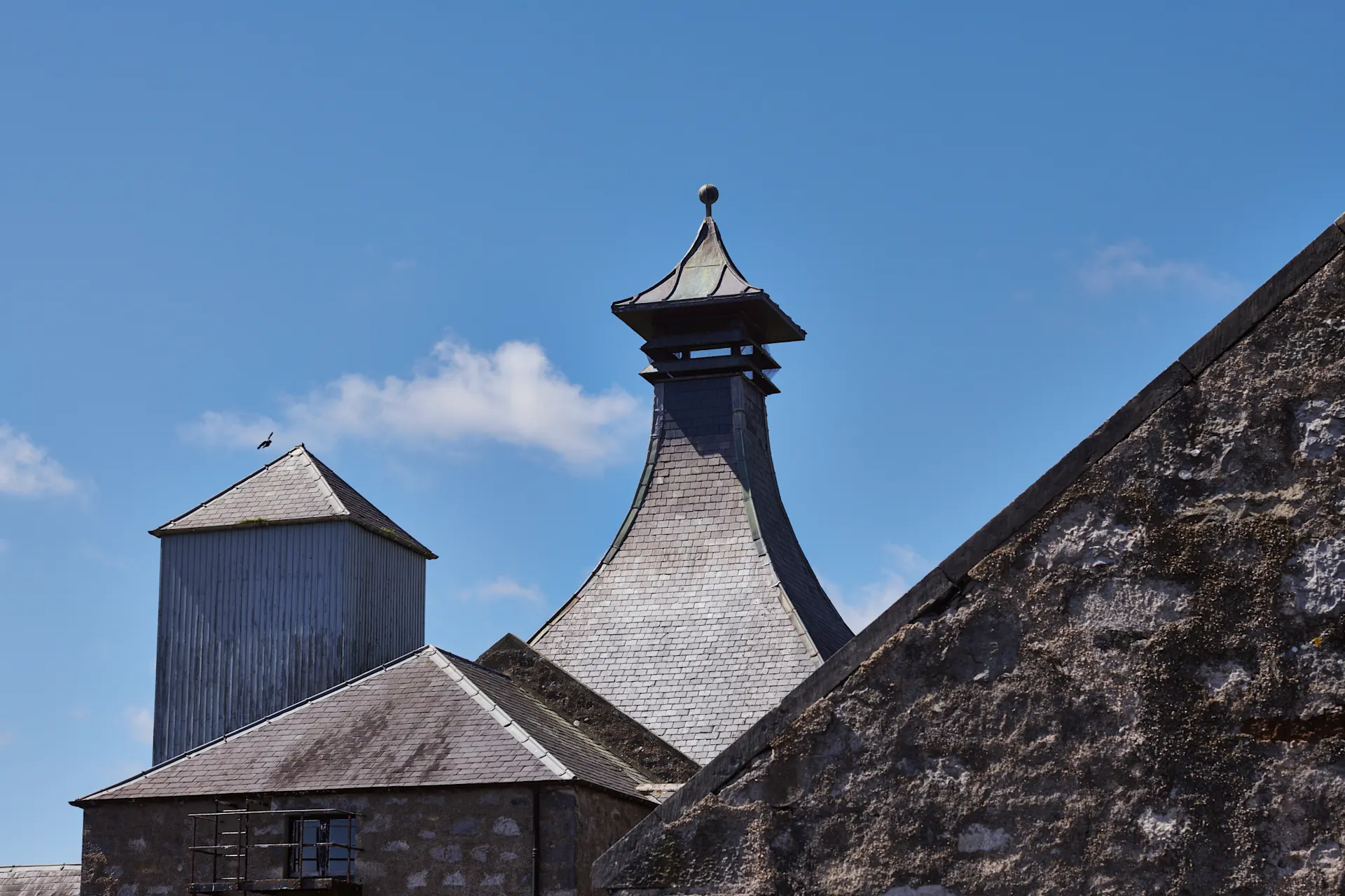 Het schuine dak van de Brora distilleerderij is te zien tegen een helderblauwe lucht, omringd door de andere gebouwen van de distilleerderij.