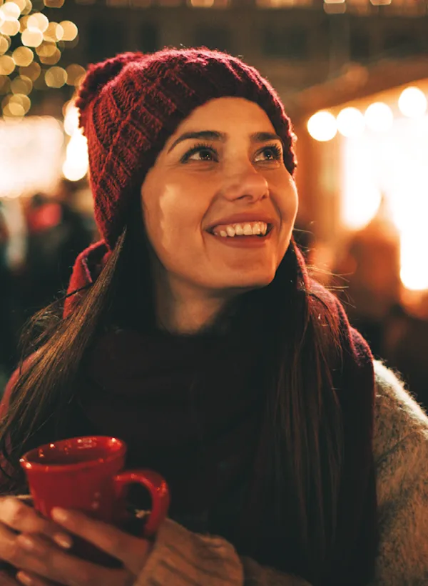A smiley woman holding a red mug looking to the horizon with Christmas light in the background