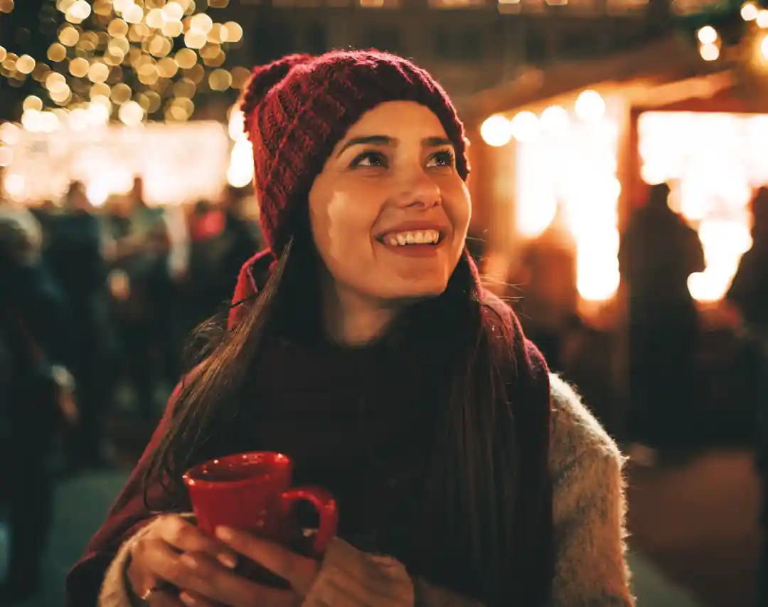 A smiley woman holding a red mug looking to the horizon with Christmas light in the background