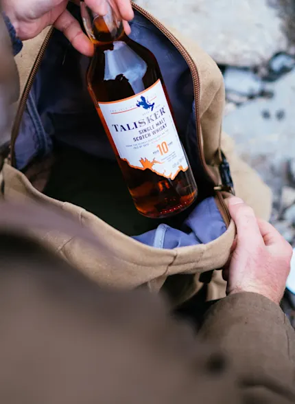 Eine Person nimmt eine Flasche Talisker 10 Jahre alten Whisky aus einem Rucksack am Meer