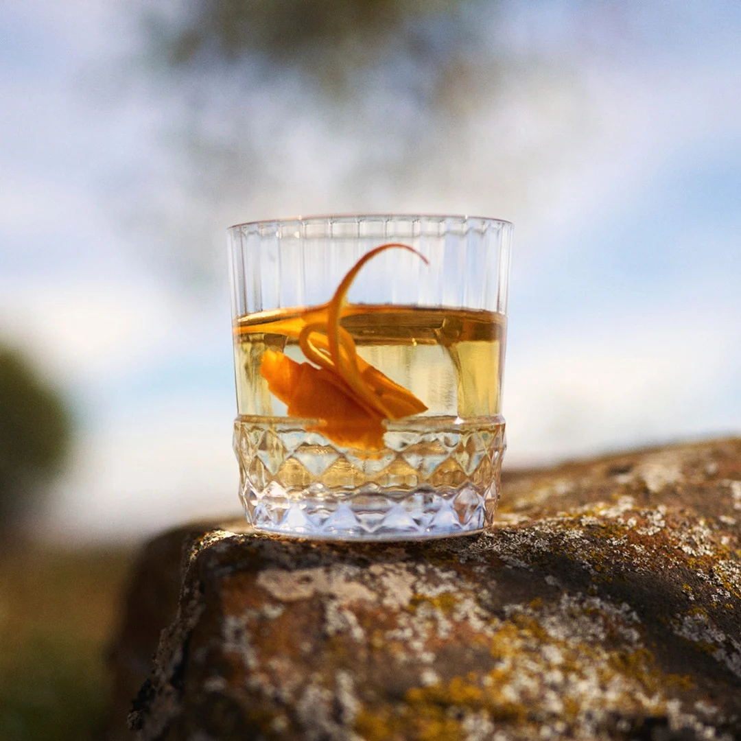 An Old Fashioned with an orange garnish on a rock in the Scottish countryside.