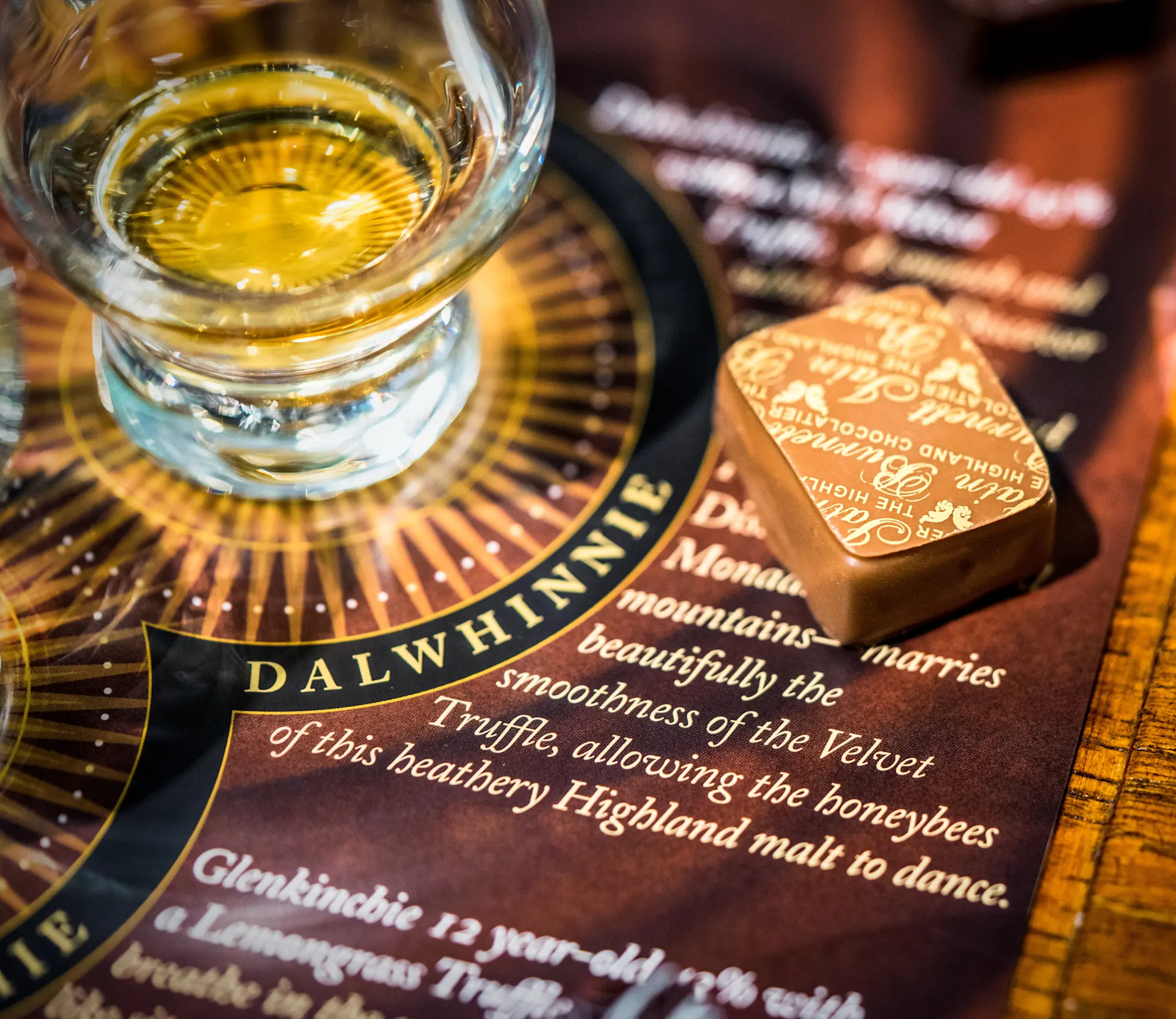 Ein Stück Schokolade liegt auf einem Stück Papier mit der Aufschrift Dalwhinnie, daneben ein Glas Whisky.