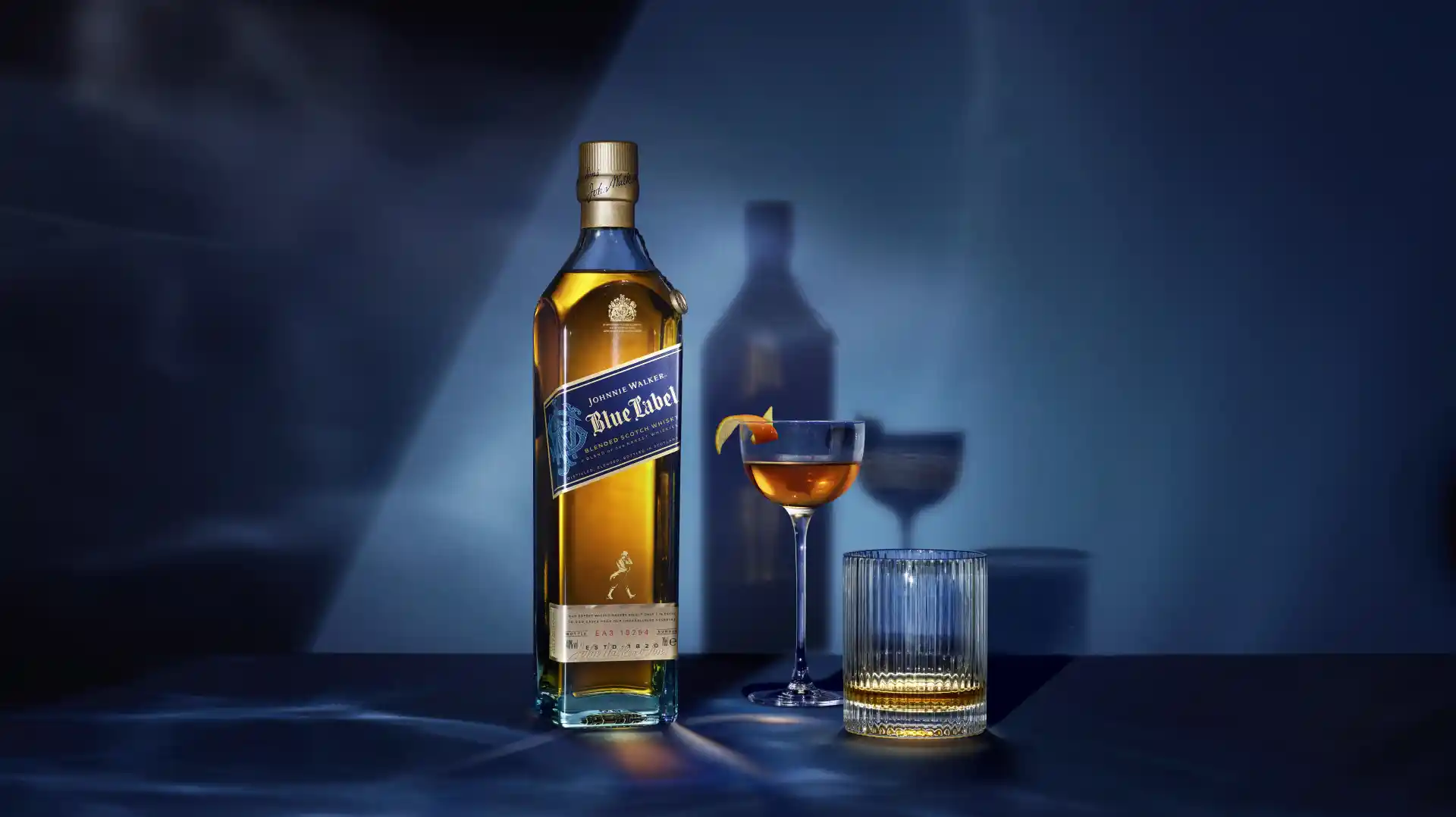 A bottle of Johnnie Walker Blue Label stands on a blue surface against a blue background, with a coupe glass cocktail and a rocks glass, both containing whisky