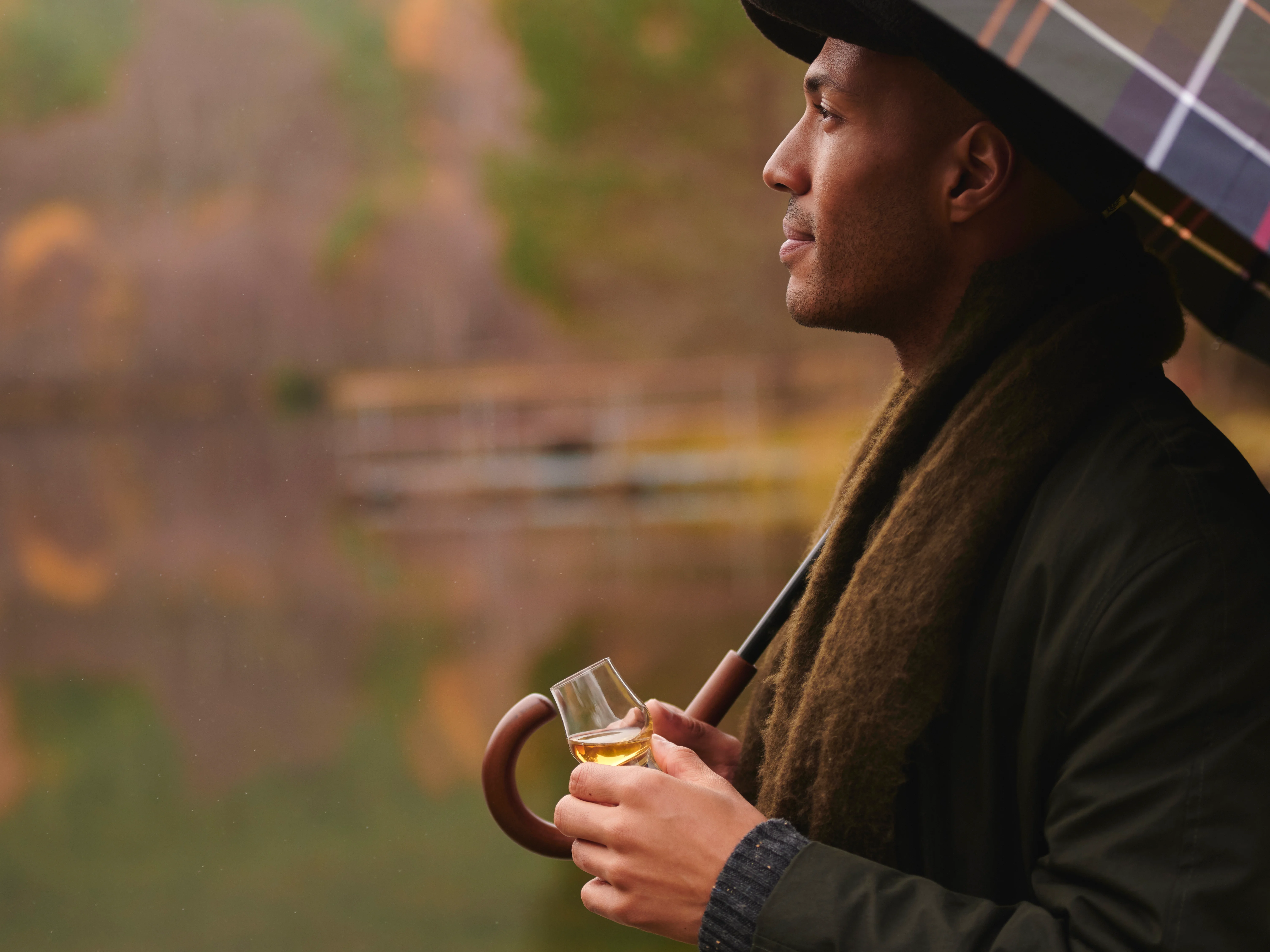 A man holds a small tasting glass of whisky while holding an umbrella over himself and standing next to a loch