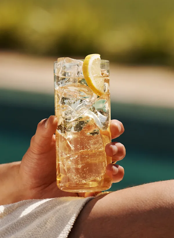 Een man houdt een whisky highball op zijn knie terwijl hij naast een zwembad zit