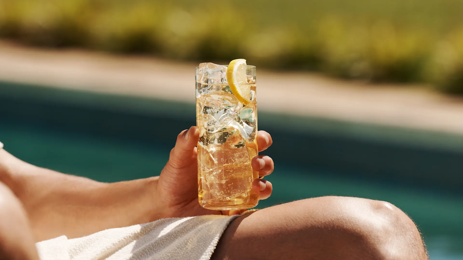 Een man houdt een whisky highball op zijn knie terwijl hij naast een zwembad zit
