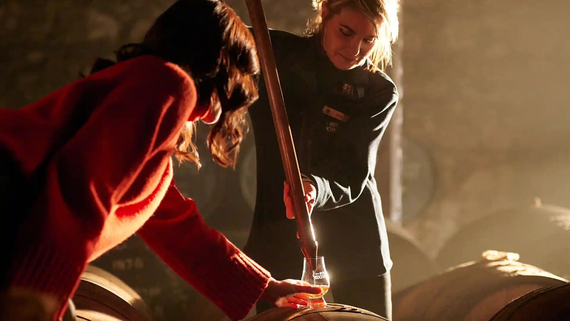 A woman extracts whisky from a barrel using a large metal pipette. She places it into a glass, which sits on top of the barrel and is being held by a woman.