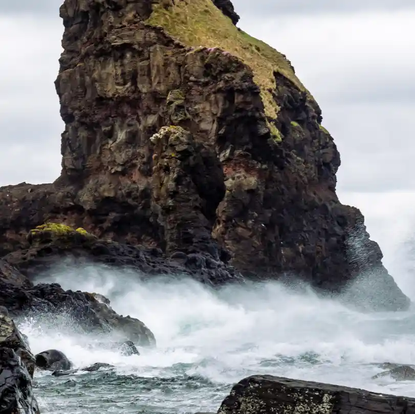 : Eine große, felsige Klippe ist von kleineren Klippen und Felsen umgeben. Ein wildes Meer prallt gegen den Fuß der Klippe und schickt Gischt in die Luft.