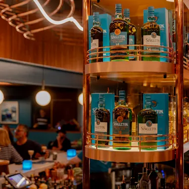 A bar with 6 bottles of Singleton on the back shelf  and people sat at the bar drinking cocktails