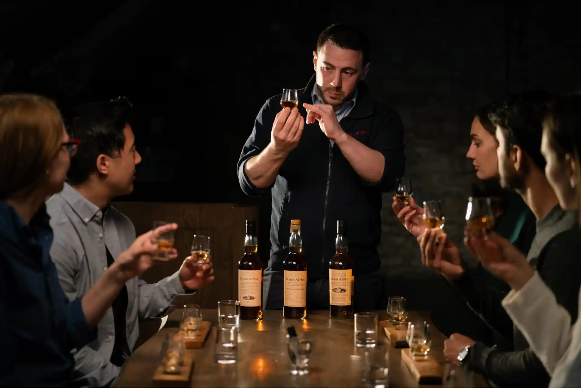 An expert leads a whisky tasting session for a group. He stands at the head of the table while they are seated, and he points to a small dram of whisky he holds. The people hold their own drams.