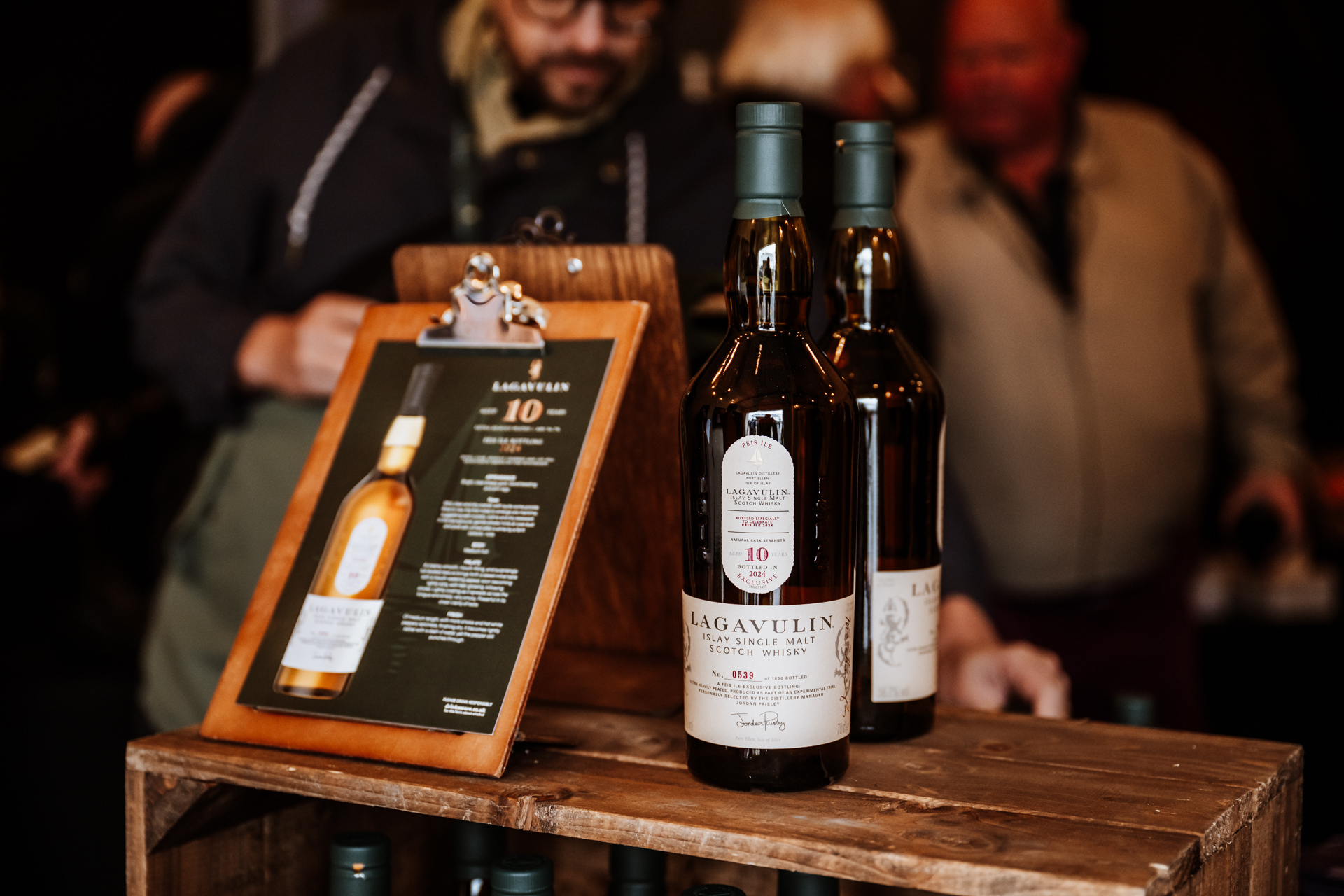 Nahaufnahme von Lagavulin 10 Jahre Islay Single Malt Scotch Whisky auf einem Holztisch während des Feis Ile Festivals. Im Hintergrund stehen Personen an einem Verkaufsstand.