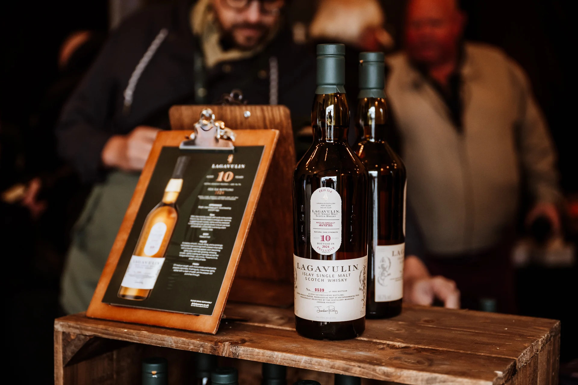 Nahaufnahme von Lagavulin 10 Jahre Islay Single Malt Scotch Whisky auf einem Holztisch während des Feis Ile Festivals. Im Hintergrund stehen Personen an einem Verkaufsstand.