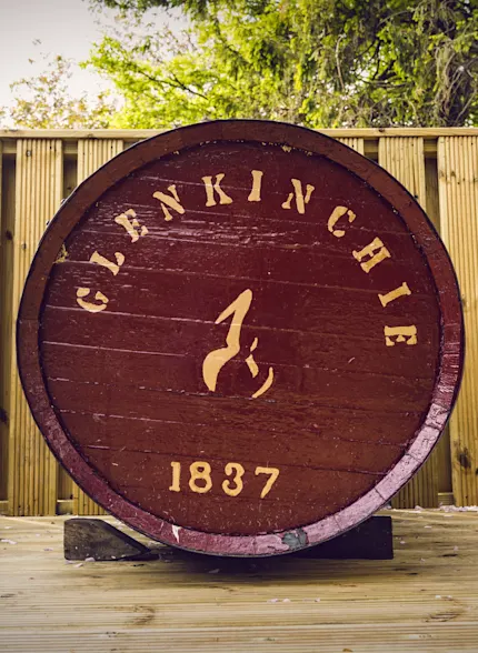 A red wooden barrel lays on its side with the front facing forwards. On the front is printed, in gold writing, ‘Glenkinchie 1837’. The barrel lays outside on a wooden surface, with a wooden fence behind it.
