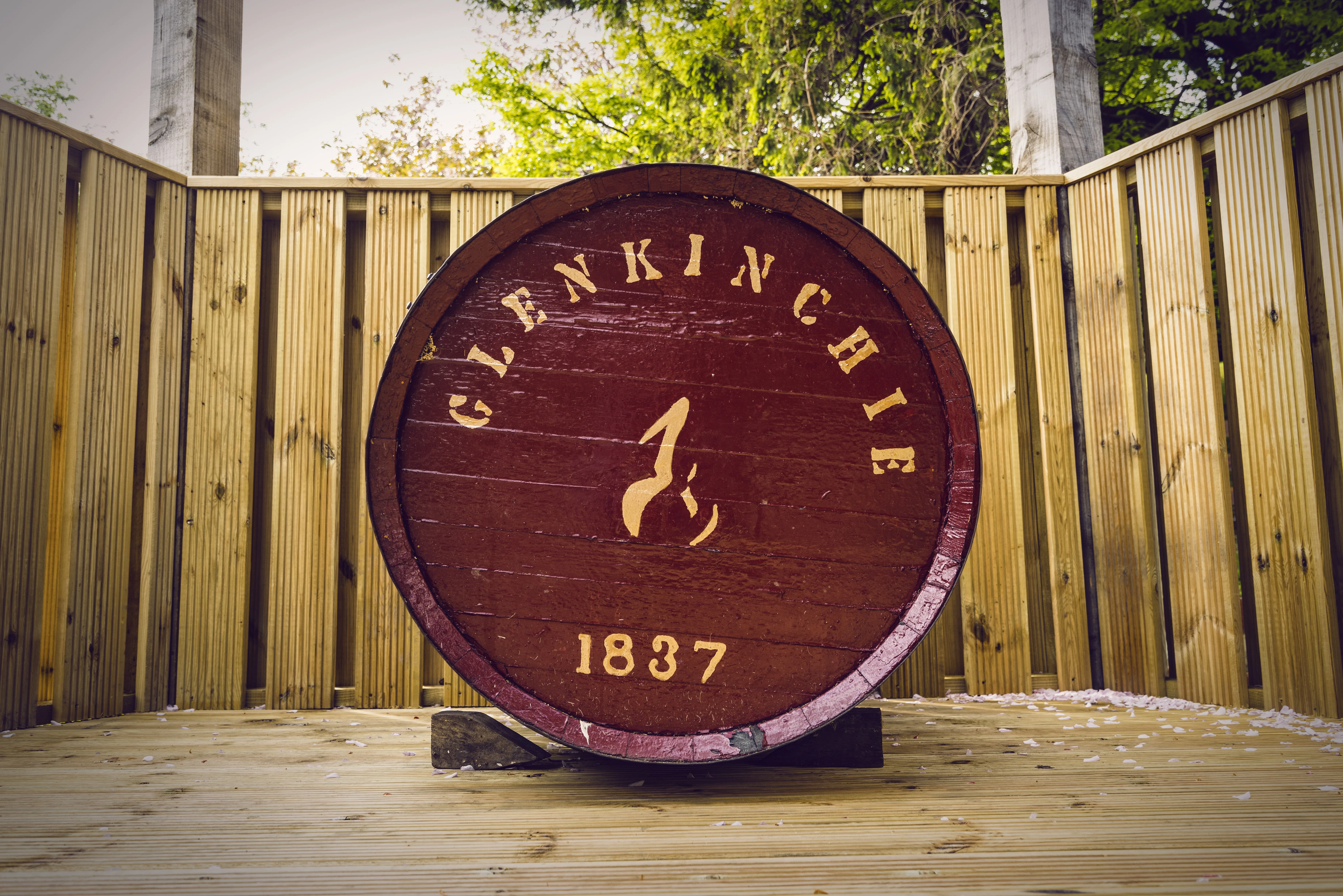 A red wooden barrel lays on its side with the front facing forwards. On the front is printed, in gold writing, ‘Glenkinchie 1837’. The barrel lays outside on a wooden surface, with a wooden fence behind it.