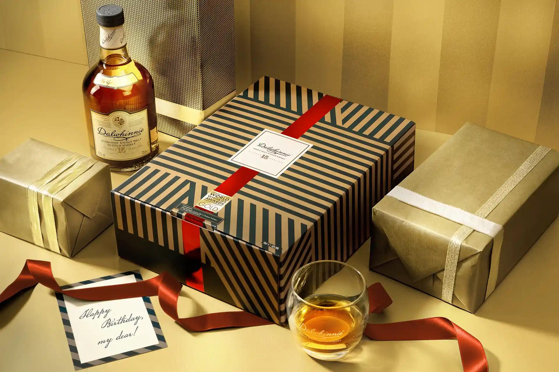 Een geschenkarrangement met een fles Dalwhinnie 15-Year-Old Single Malt Scotch Whisky, een gestreepte geschenkdoos met een rood lint, extra ingepakte cadeautjes en een glas whisky.