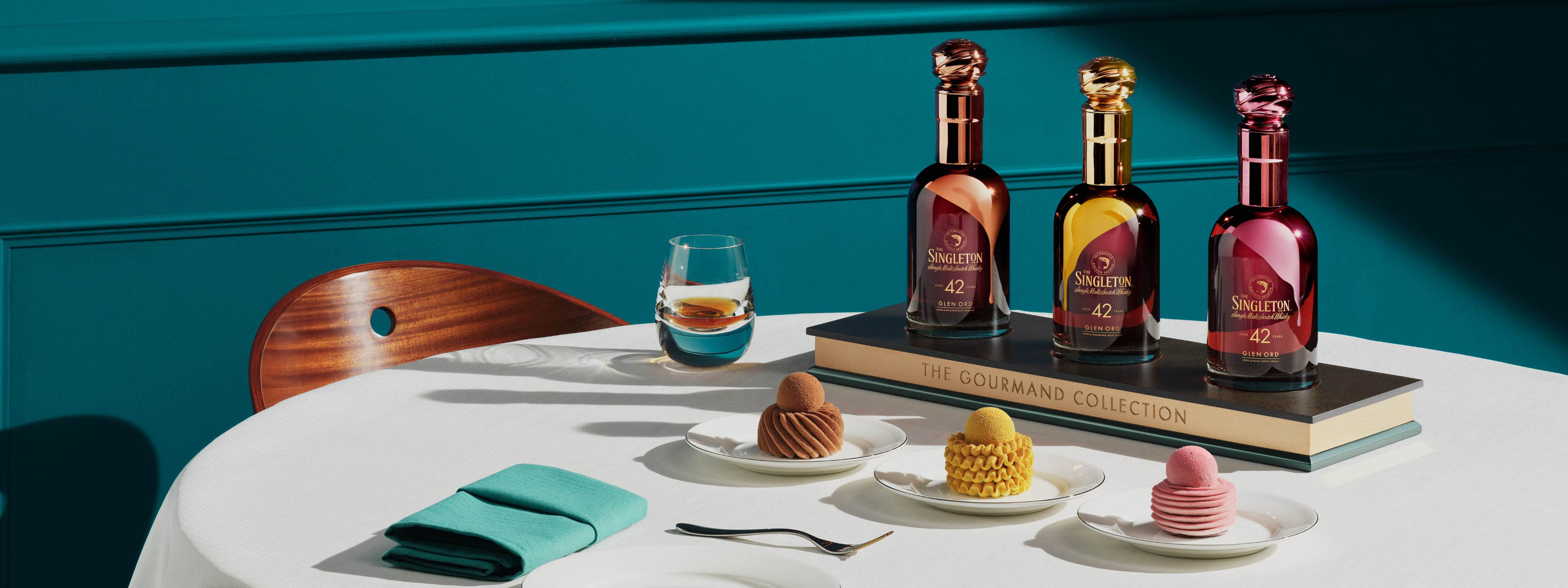 Drei Flaschen The Singleton Gourmand Collection auf einem Buch, dazu elegante Desserts und ein Glas Whiskey auf einem Tisch mit blaugrünen Akzenten.
