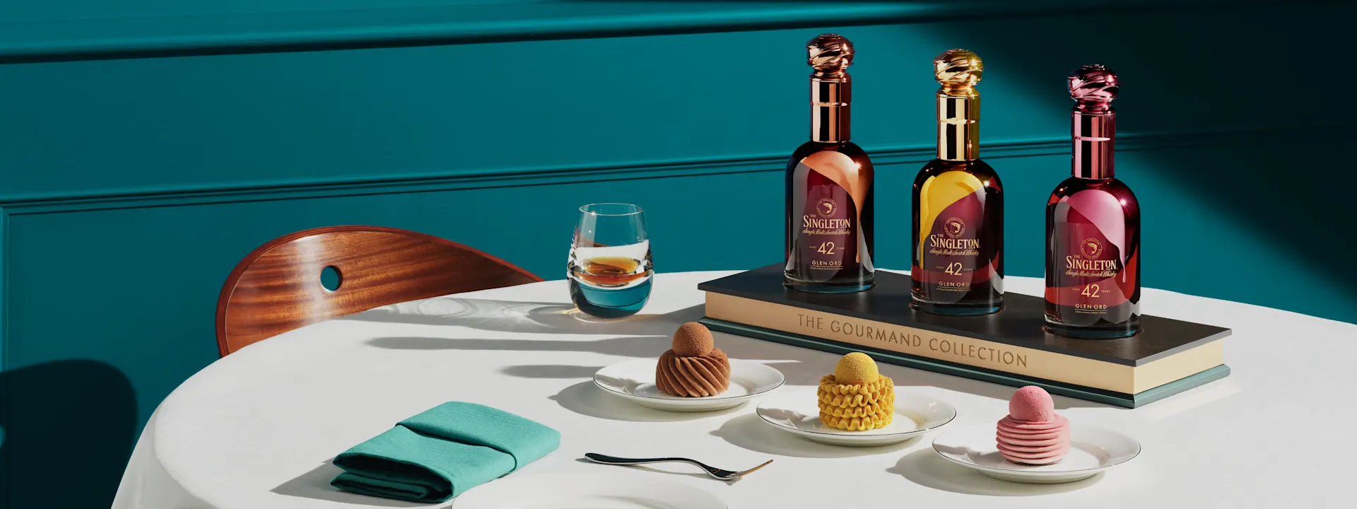  Three bottles from The Singleton Gourmand Collection displayed on a book, alongside elegant desserts and a glass of whiskey on a table with teal accents.