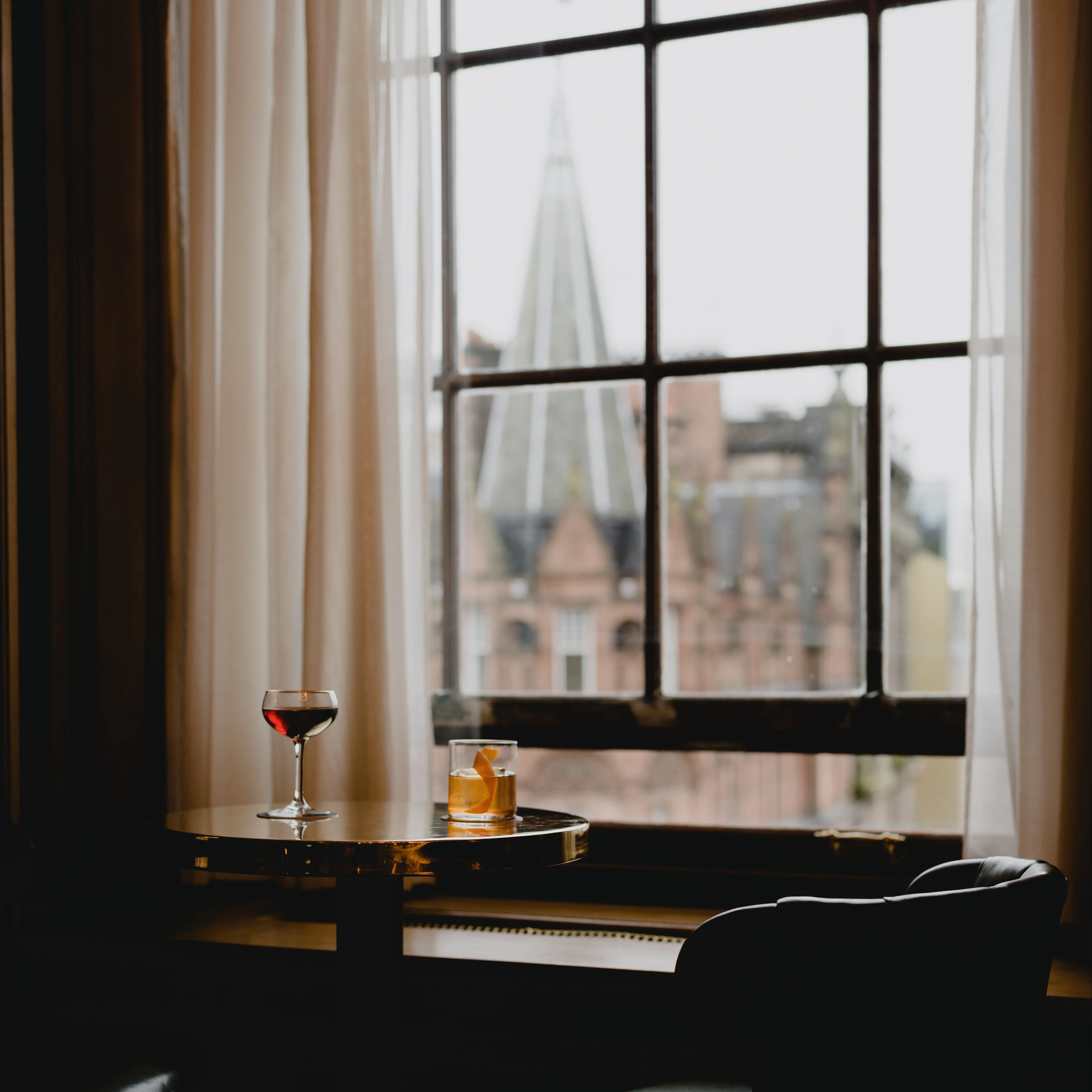 A glass of whisky and red wine on a table in front of a large bay window, filled with light.