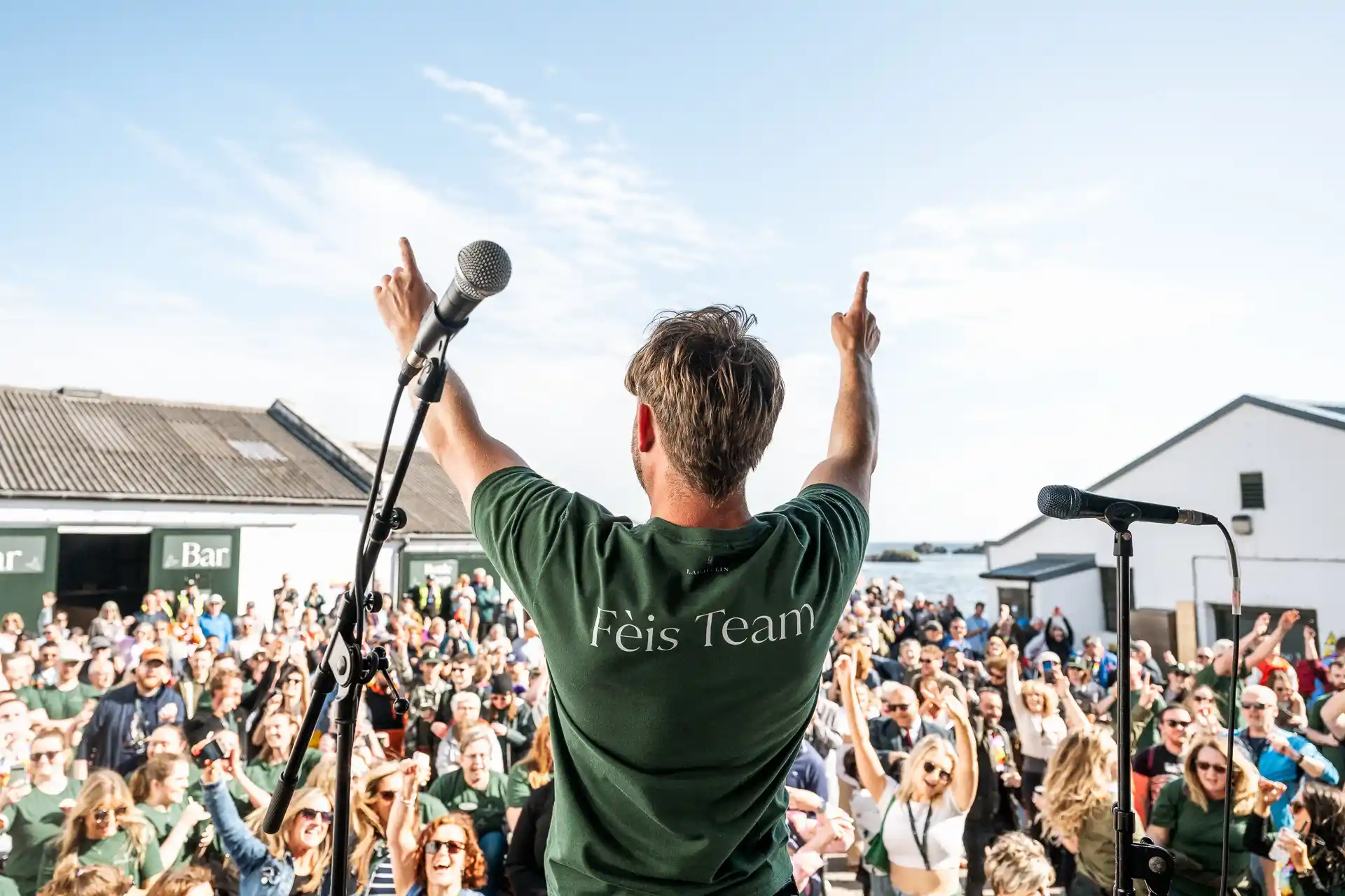 Man staat op het podium in een groen shirt op het Féis Ile festival om het publiek op te vrolijken.