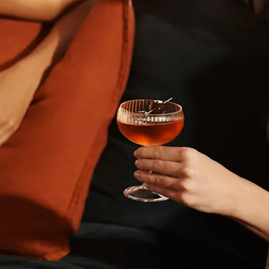 A woman holds a manhattan cocktail by the stem of the glass while sitting on a sofa with a friend