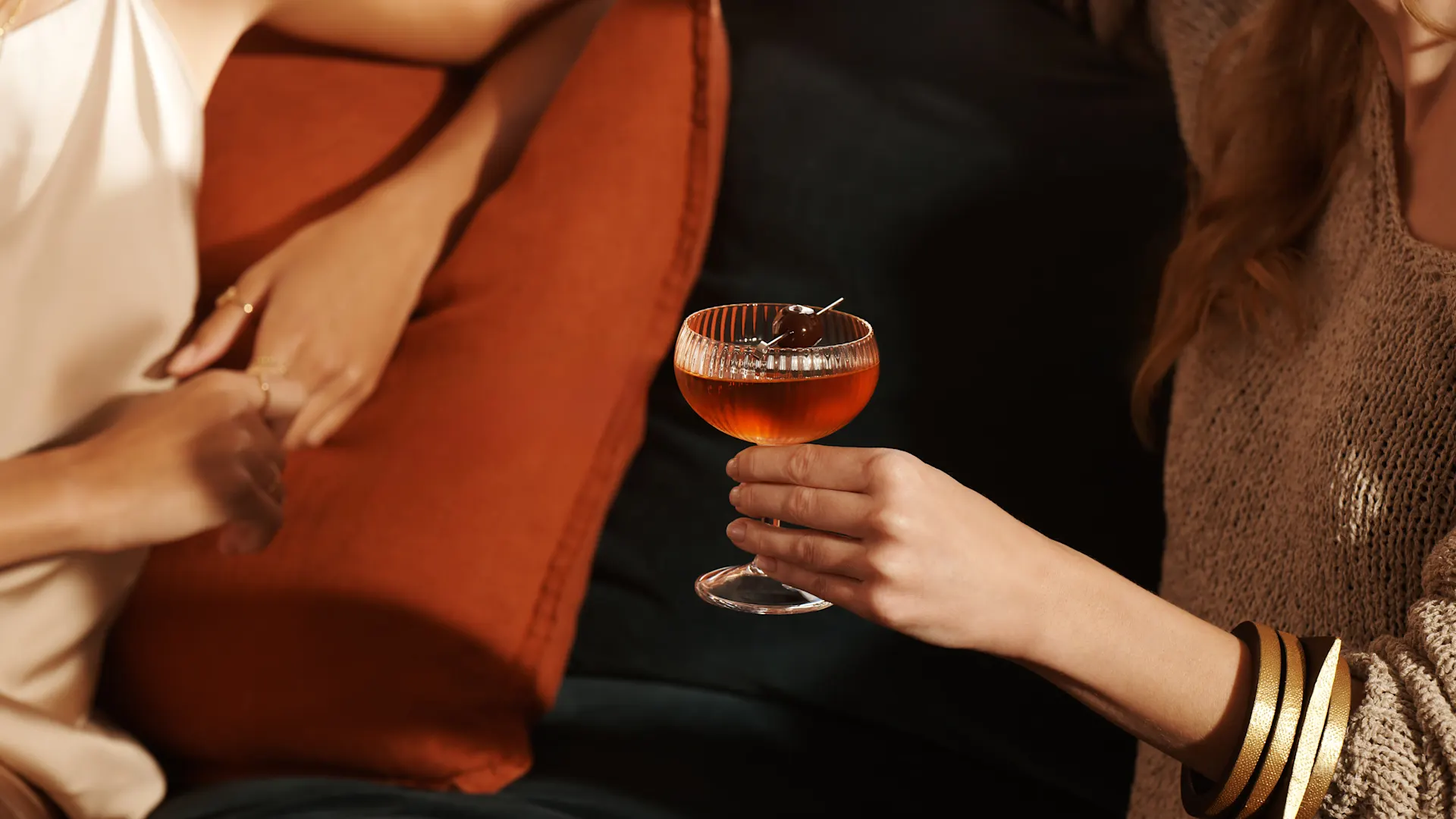 Een vrouw houdt een Manhattan-cocktail vast bij de steel van het glas terwijl ze met een vriendin op een bank zit