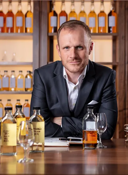 Dr Craig Wilson sits behind a wooden table laden with glass bottles containing various brown liquids, books and a notepad and pen. He wears a suit and looks at the camera. Behind him are display shelves holding many more bottles of brown liquids.