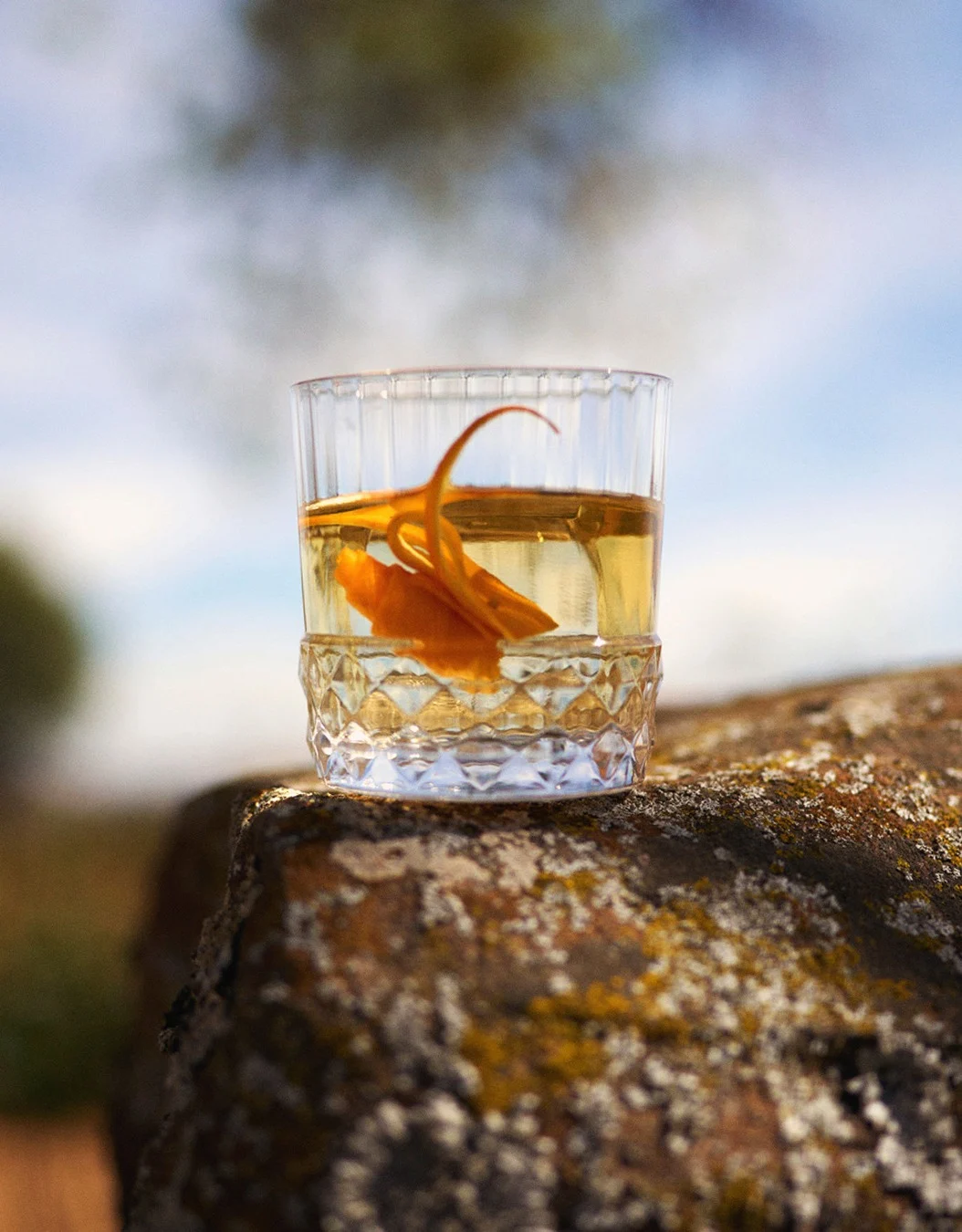 A whisky cocktail in a rocks glass on a rock in the countryside. 