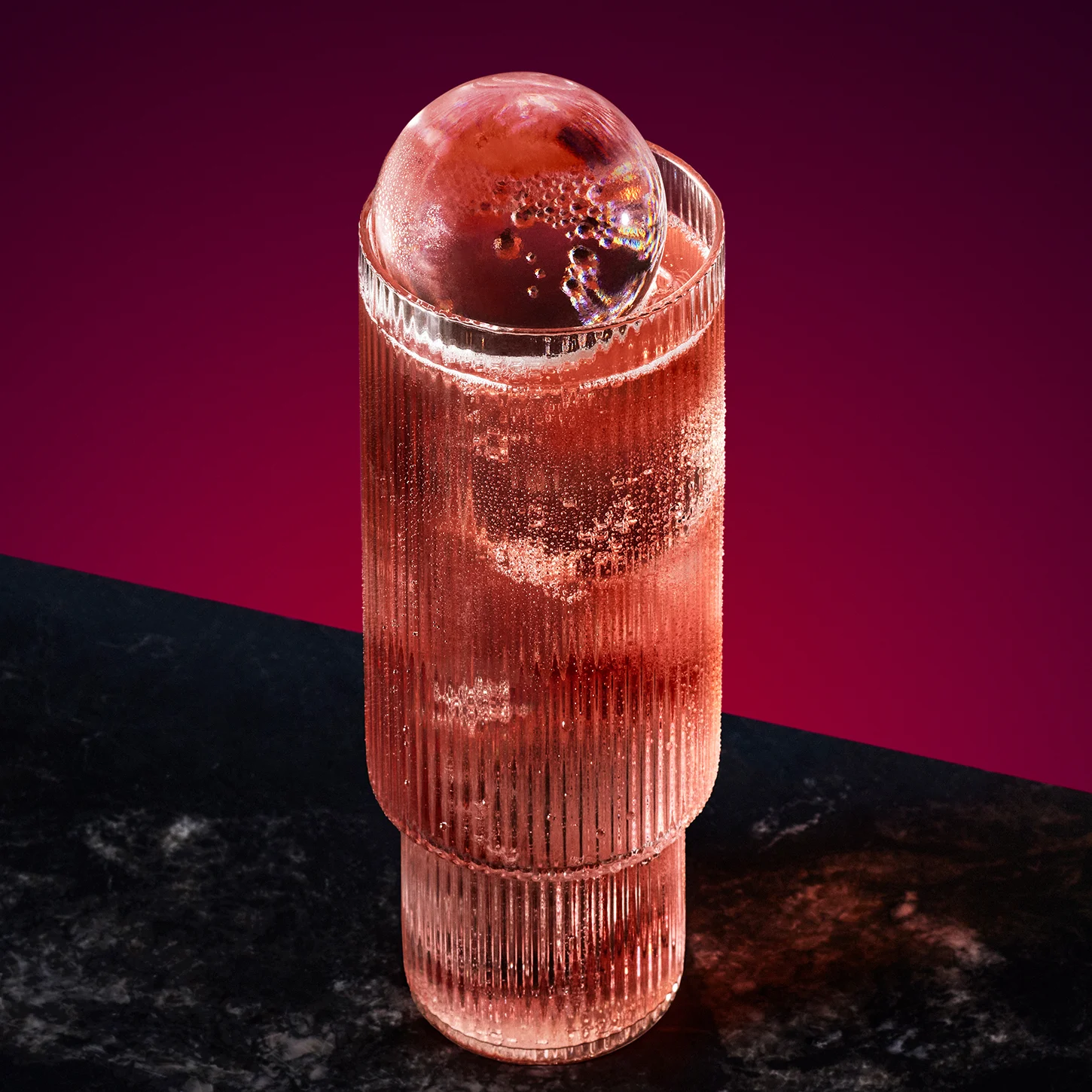 Tall glass of chilled pink drink with a large spherical ice cube on top, placed against a gradient purple background. The drink is placed on a dark surface, with the light casting a subtle shadow.