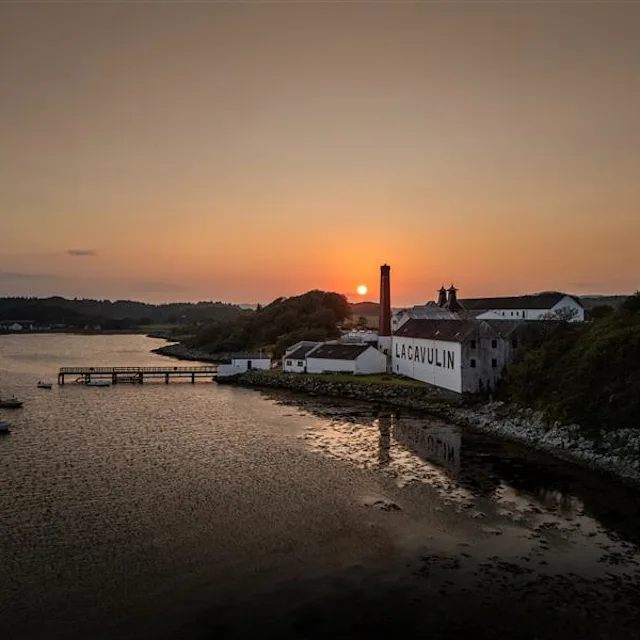 A woman uses a large pipette to take whisky from a wooden barrel and place it into a glass held by a man wearing a green Lagavulin t-shirt. Behind them sits several people and lots of other wooden barrels.