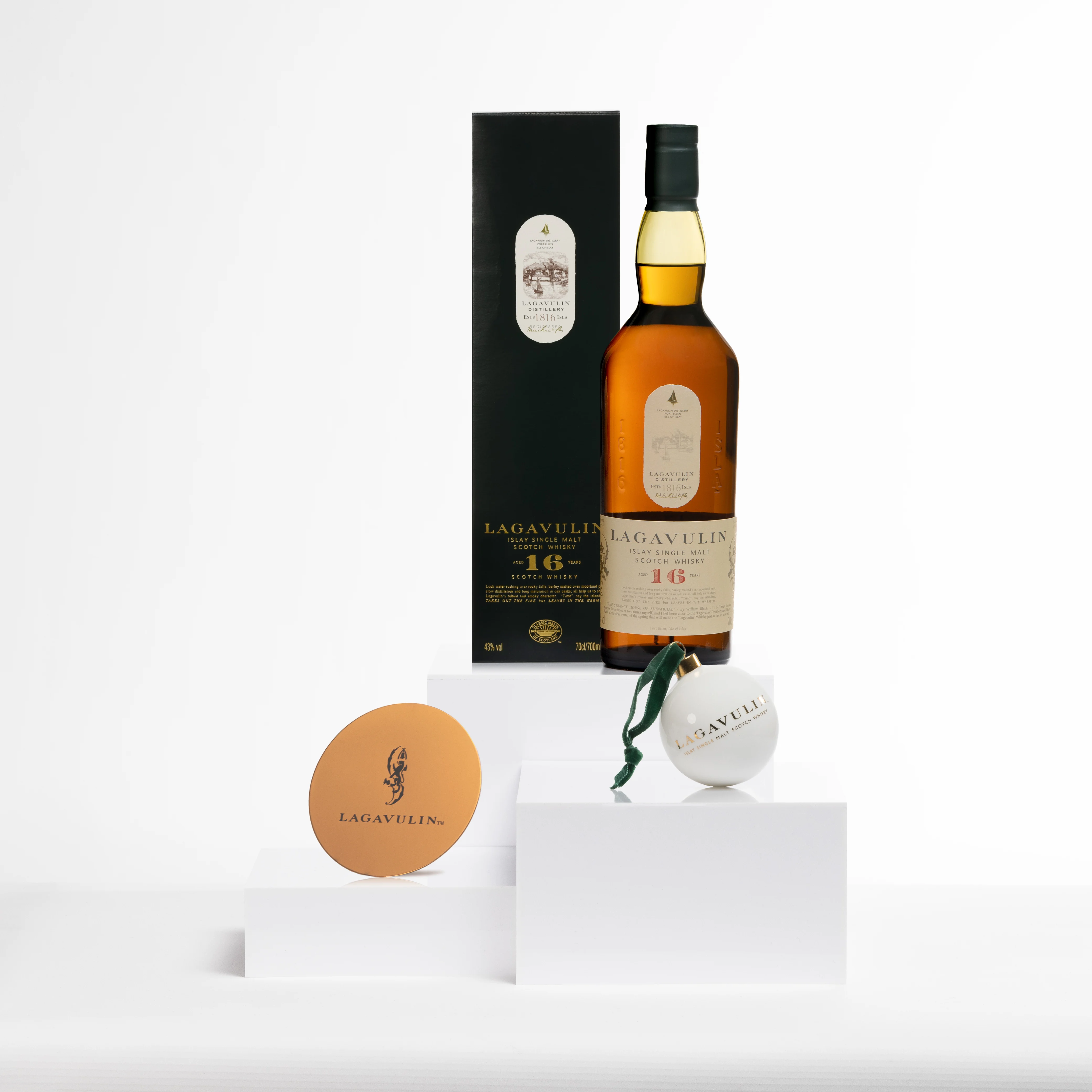 A bottle of Lagavulin 16-Year-Old Scotch whisky, a copper-toned coaster with the Lagavulin logo, and a white bauble with green ribbon branding, arranged on sleek white blocks.