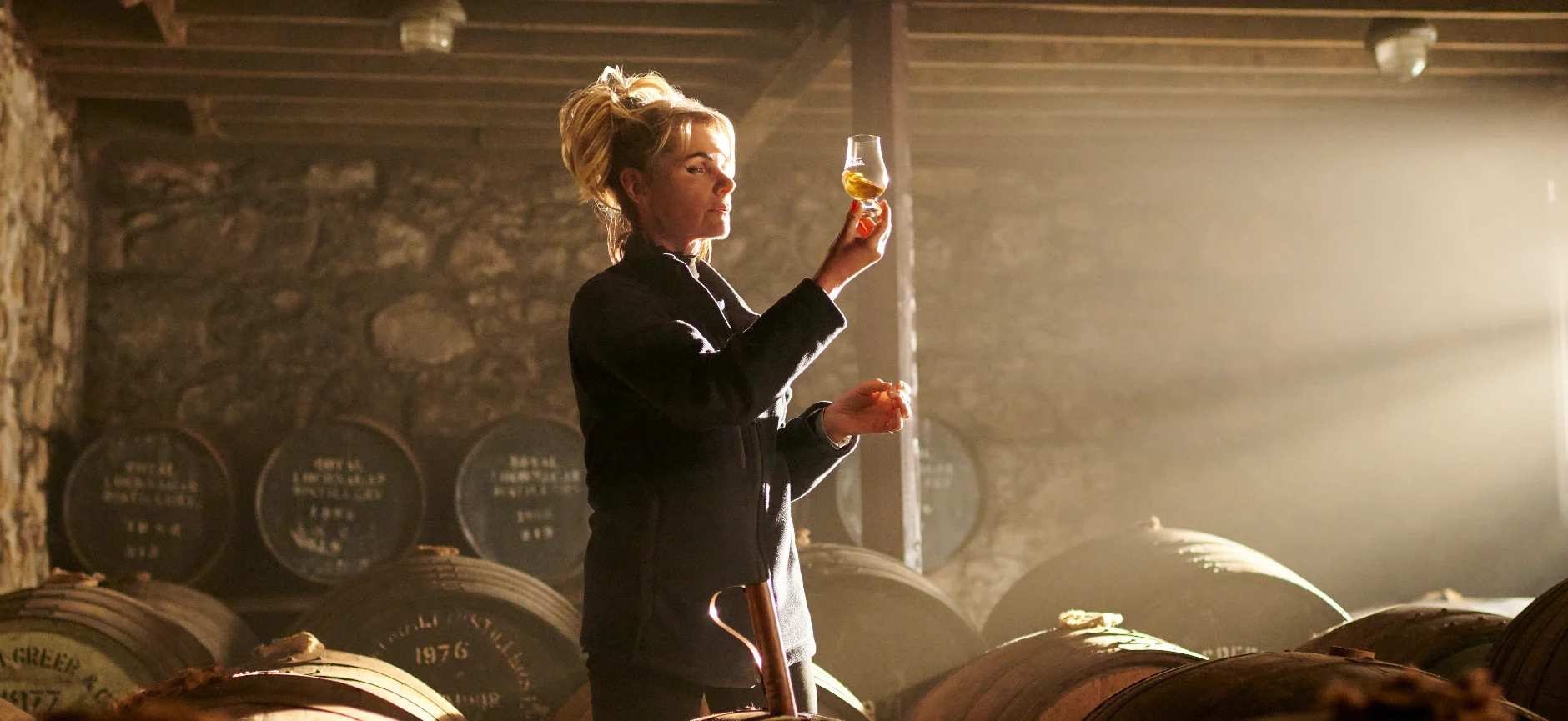 A person holding a glass of golden Scotch whisky up to the light in a Scottish Highland distillery setting, with warm amber tones highlighting the whisky's colour
