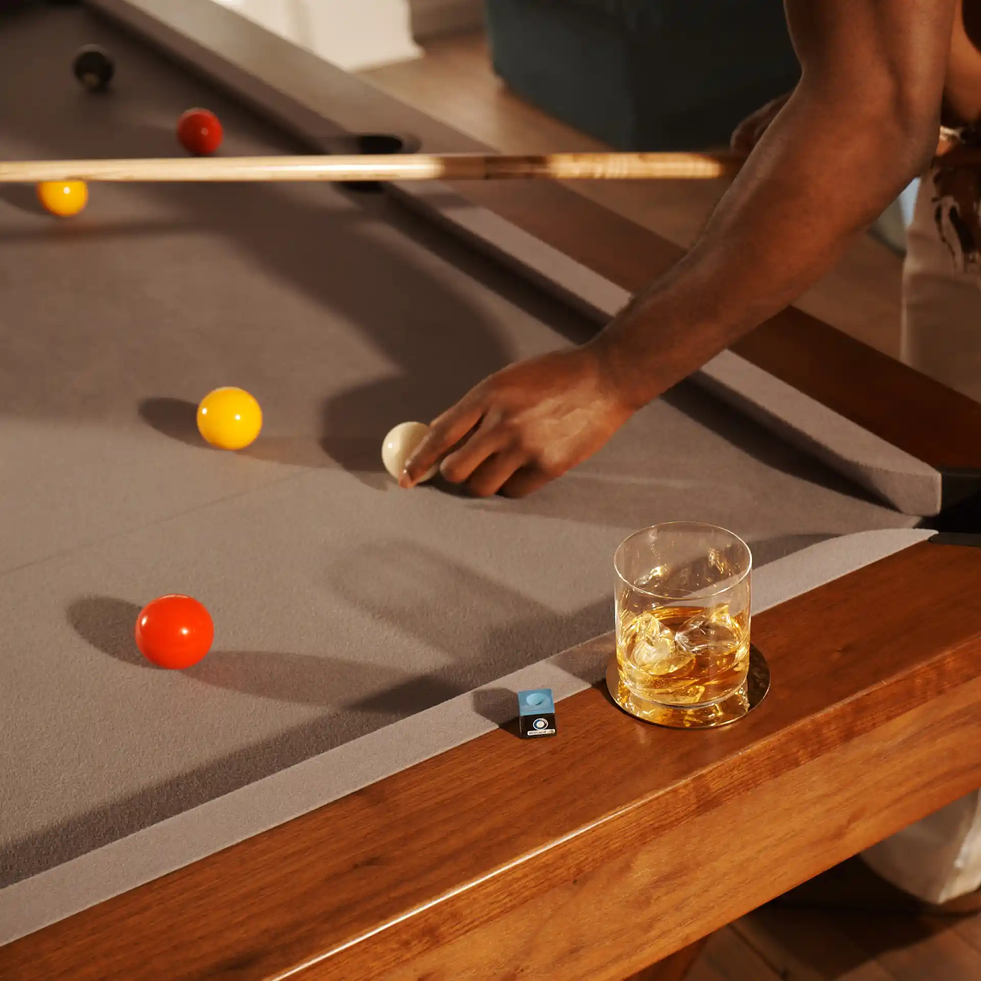 A rocks glass containing whisky and ice sits on the edge of a pool table which contains several pool balls. Someone stands over the table, holding a cue and repositioning a white ball.