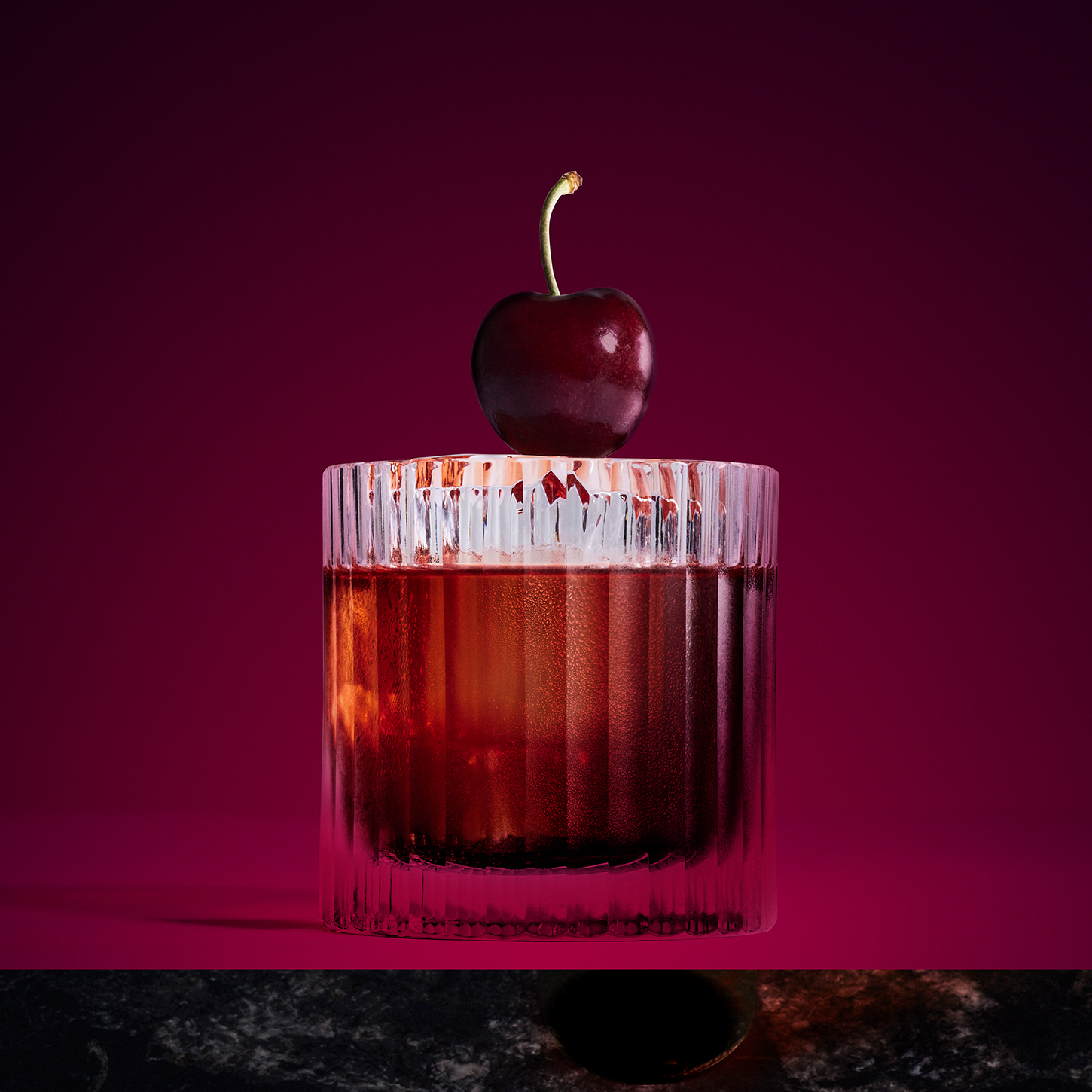 A glass of whiskey with a clear ice cube, garnished with a cherry on the rim, sits on a dark marble surface against a deep purple background. Shadows create a dramatic contrast.