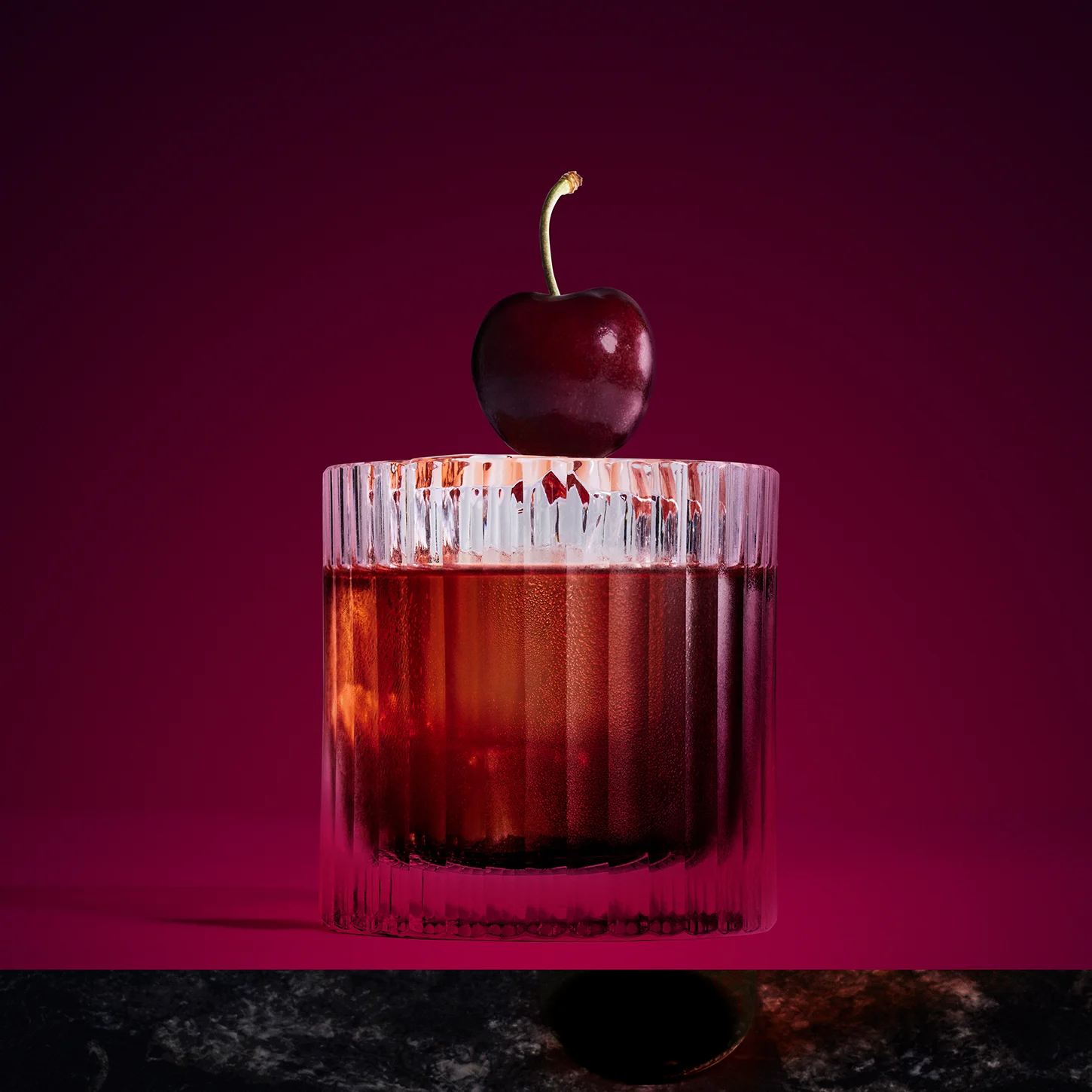 A glass of whiskey with a clear ice cube, garnished with a cherry on the rim, sits on a dark marble surface against a deep purple background. Shadows create a dramatic contrast.