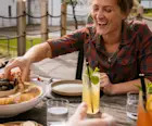 Katie Tunn, a blonde woman, sits outside at a wooden table laden with plates and bowls of food. She reaches for food on a plate while smiling.