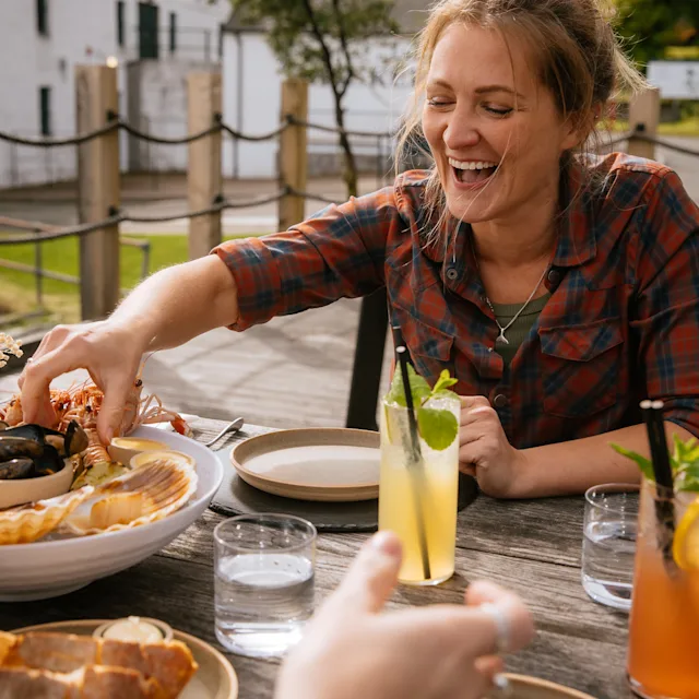 Katie Tunn, een blonde vrouw, zit buiten aan een houten tafel vol met borden en schalen met eten. Ze reikt glimlachend naar het eten op een bord.