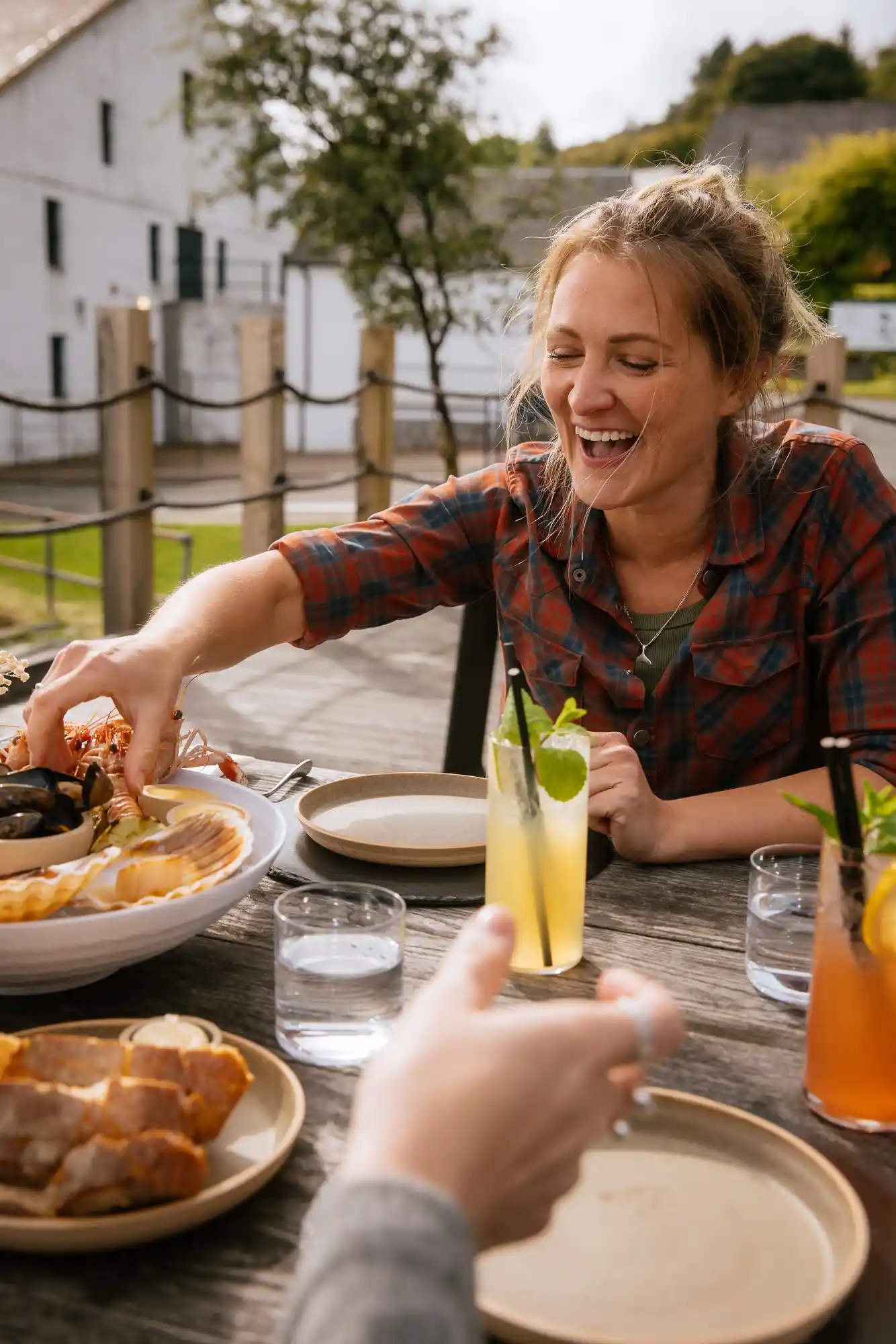 Katie Tunn, a blonde woman, sits outside at a wooden table laden with plates and bowls of food. She reaches for food on a plate while smiling.