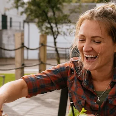 Katie Tunn, een blonde vrouw, zit buiten aan een houten tafel vol met borden en schalen met eten. Ze reikt glimlachend naar het eten op een bord.